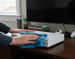 A person cleaning the exterior of an Xbox One S console using a microfiber cloth
